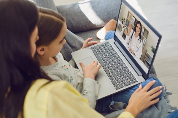 Mamma och barn sitter med laptop där en läkare visas på skärmen