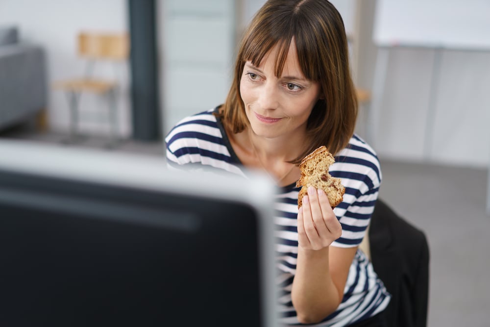 Office Woman Holding a Bread Snack While Working on her Computer at her Table.