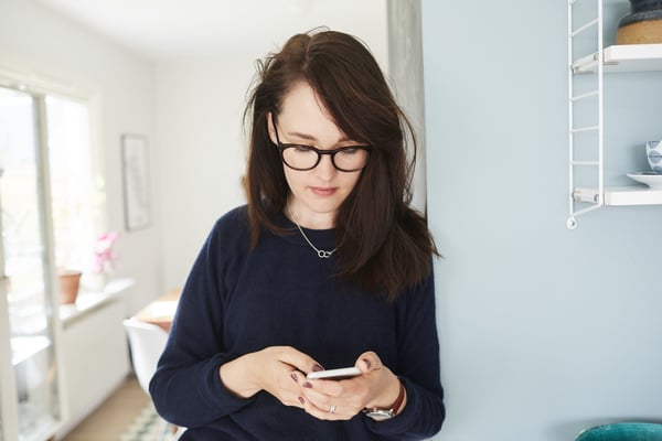 Ung kvinna står i sitt hem och tittar ner i mobilen
